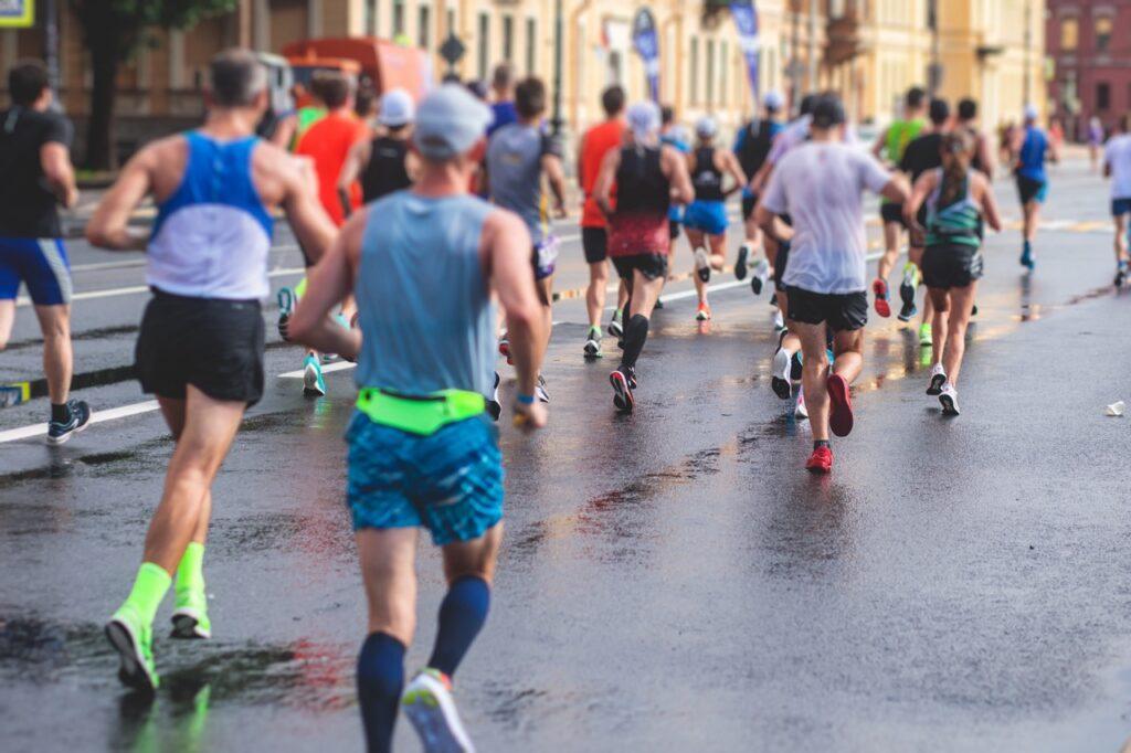 Bildet viser en gruppe løpere som deltar i et maraton eller løp på en bygate. Det er en blanding av menn og kvinner, alle kledd i treningstøy og joggesko. Gaten er fuktig, som om det nylig har regnet, og reflekterer fargene fra løpernes klær og sko. Bygninger og andre løpere kan sees i bakgrunnen, men fokus er på ryggen til løperne, som er i ferd med å løpe videre mot mål. Løperne på bildet har på seg typisk løpeutstyr: lette løpeshorts, tekniske t-skjorter eller singlets, løpesokker, og spesialiserte joggesko for komfort og ytelse. Noen har også capser eller kompresjonsplagg for ekstra støtte. Vi gir deg 8 morsomme fakta om maraton!