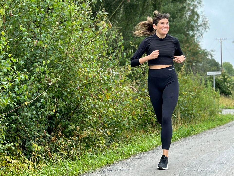 Bildet viser en kvinne som jogger langs en vei omgitt av grønn, frodig vegetasjon. Hun er kledd i svarte treningstights og en svart langermet topp, og smiler bredt mens hun løper. Hun ser avslappet og glad ut, og bildet formidler en følelse av glede og energi knyttet til løping i naturen. Bakgrunnen viser høye trær og busker, og det ser ut til å være en overskyet dag, men stemningen er likevel positiv og oppfriskende. Bildet understreker viktigheten av å smile og ha det gøy mens man trener. Vi forklarer hvorfor du bør smile når du trener, og hva som da skjer!