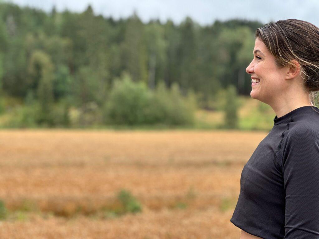 Bildet viser en kvinne som står i profil med et smil om munnen. Hun har på seg en svart treningsgenser med lange ermer. I bakgrunnen er det et åpent landskap med høstfargede åkre og skogkledde trær, som er litt uskarpe på grunn av dybdeskarpheten. Været ser ut til å være overskyet, og det skaper en myk og nøytral atmosfære i bildet. Kvinnen ser ut til å være ute på en joggetur eller trening i naturen, og smilende uttrykker hun glede på grunn av en følelse av "runner's high". Vi forteller hva runners high er, når det kommer, hvorfor det kommer og om alle får det!