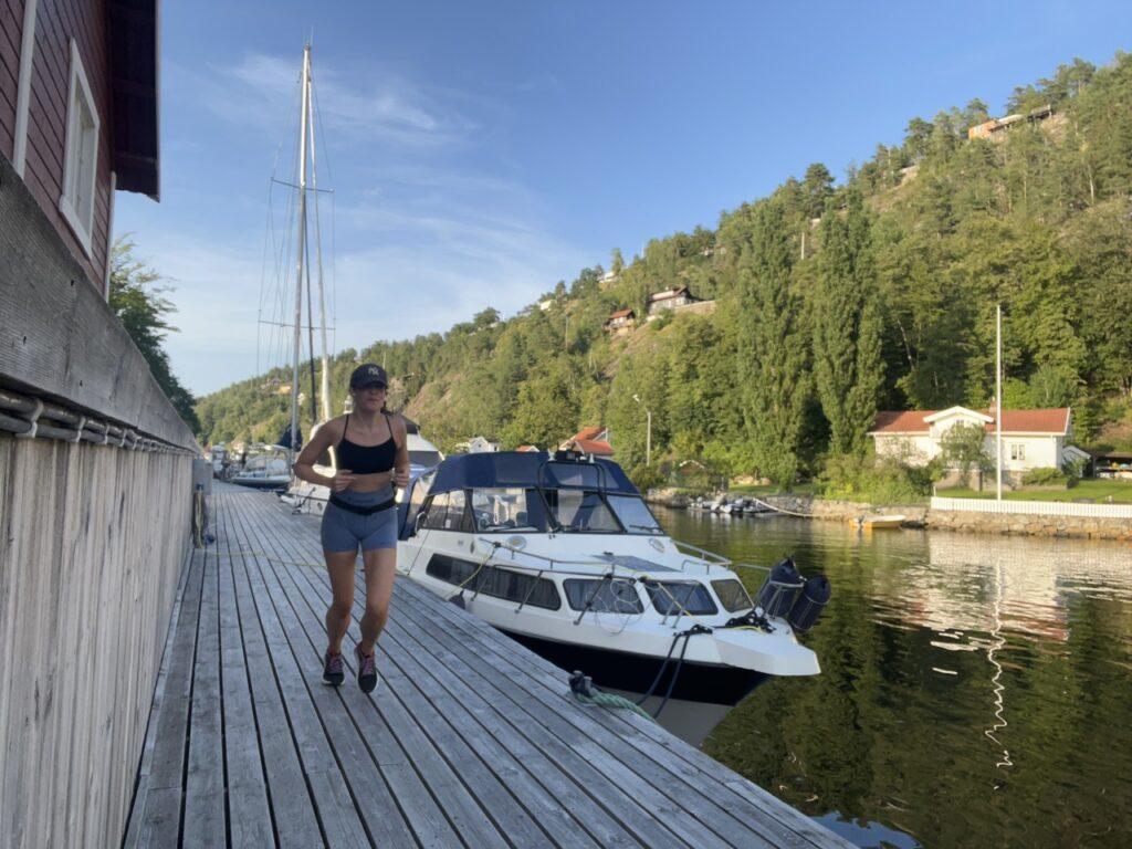 Bildet viser en kvinne som løper langs en brygge ved en marina, omgitt av båter som ligger til kai. Hun har på seg en mørk NY-yankees caps med "NY"-logo, en sports-BH og korte treningsshorts. I bakgrunnen kan man se en rekke båter langs vannet og en grønn åsside med hus som ligger spredt oppover åsen. Solen er lavt på himmelen, noe som gir en varm og gyllen belysning over landskapet. Vannet er rolig, og det skaper refleksjoner av båtene og vegetasjonen rundt. Bildet formidler en følelse av aktivitet og energi i et naturskjønt og fredelig miljø. Løpetrener og løpeekspert Andreas Gossner fra Løpetrening.no svarer på Hvordan og hvorfor løpe ganske hardt på en 10 km?