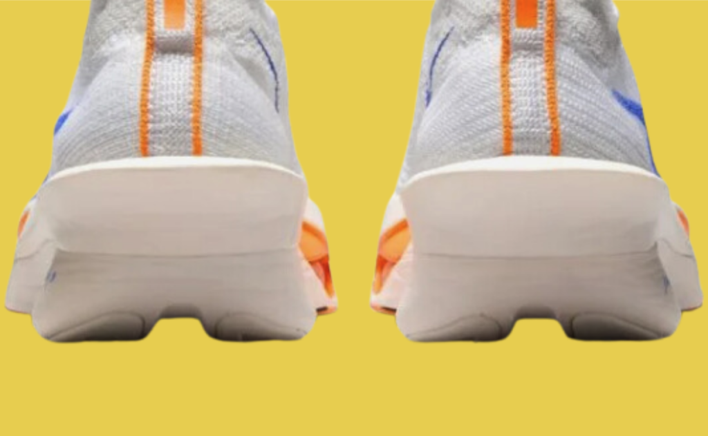 Bildet viser baksiden av et par joggesko som ser ut til å være Nike Vaporfly-modellen. Skoene har en lys grå overdel med oransje striper langs midten av hælen. Sålen er hvit med oransje innslag på sidene. Dette er en kjent modell som ofte refereres til som en av verdens raskeste løpesko, designet for løpere som ønsker maksimal ytelse. Den brede sålen og formen på hælen antyder at skoene er laget for å gi støtte og energiutnyttelse under løping.