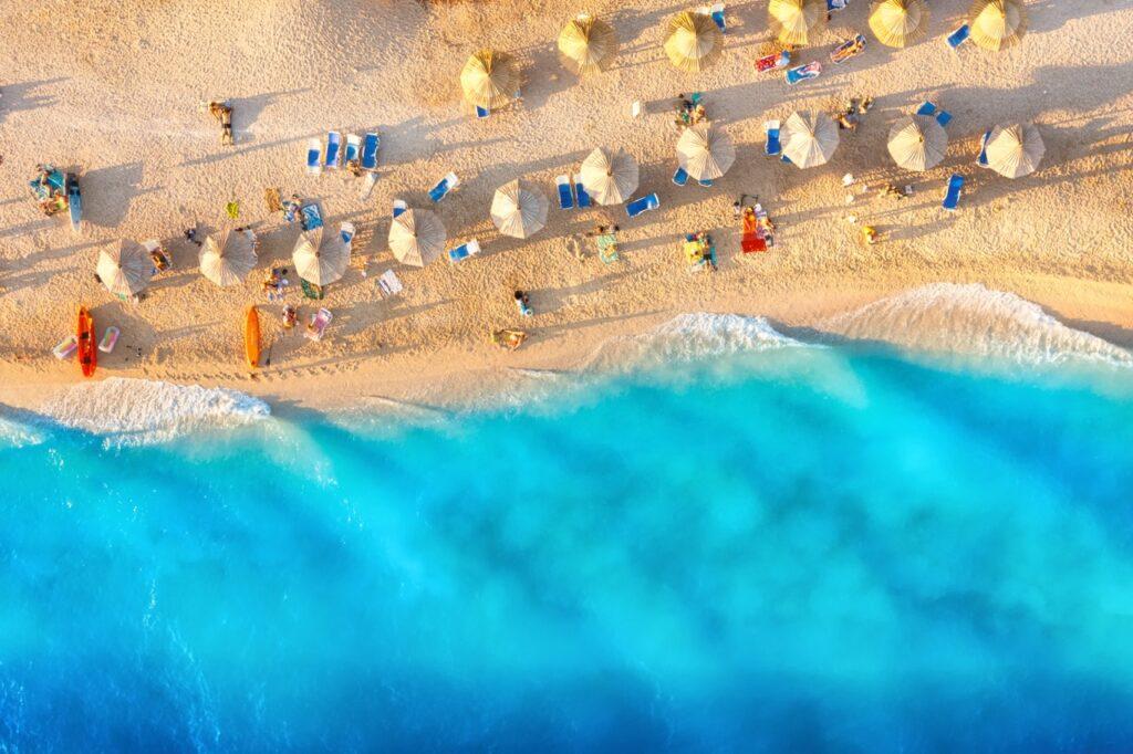 Bilde tatt ovenfra og ned av en gylden strand i Middelhavet. På stranden ser man solsenger og parasoller og azur-blått hav som skyller inn over den vakre stranden. Vi svarer på når høstferien 2024 er, samt hjelper deg med å finne billige reiser til syden i høstferien slik at du kan forlenge sommerfølelsen.