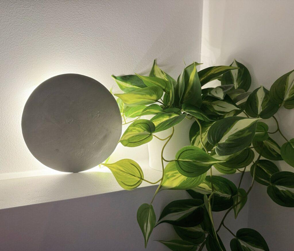 Bildet viser en rund lampe laget av selvtørkende leire som står på en hylle. Lampen er opplyst bakfra med et LED-lys, som skaper en varm glød mot veggen. Ved siden av lampen er en frodig grønn plante med store blader, som gir en naturlig og dekorativ kontrast til den minimalistiske leirelampen. Sammen gir lampen og planten et moderne og avslappende preg til rommet.