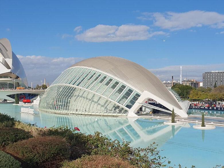 Bildet viser et futuristisk, kuppelformet byggverk som speiler seg i en stor, blå vannflate. Strukturen har en glassfasade og ligner et øye, sannsynligvis en del av Ciudad de las Artes y las Ciencias i Valencia, Spania. Bygningen står i et komplekst anlegg omgitt av vann, og bakgrunnen viser blå himmel med noen skyer. I tillegg er det busker og planter i forgrunnen, mens noen bygninger og trær kan skimtes i bakgrunnen. Arkitekturen har et moderne og futuristisk preg som står i kontrast til det naturlige landskapet rundt. Dette er løpetrenerens favorittøkt for trening til maraton!