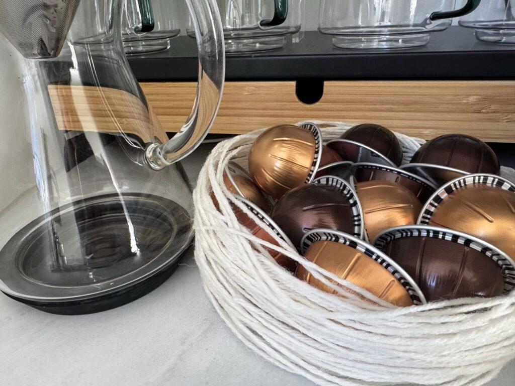 En bolle laget av hvitt garn er fylt med fargerike kaffekapsler i metalliske toner. Bollen står ved siden av en moderne glasskanne på en lys benkeplate, og gir et stilfullt og kreativt uttrykk i kjøkkeninnredningen.