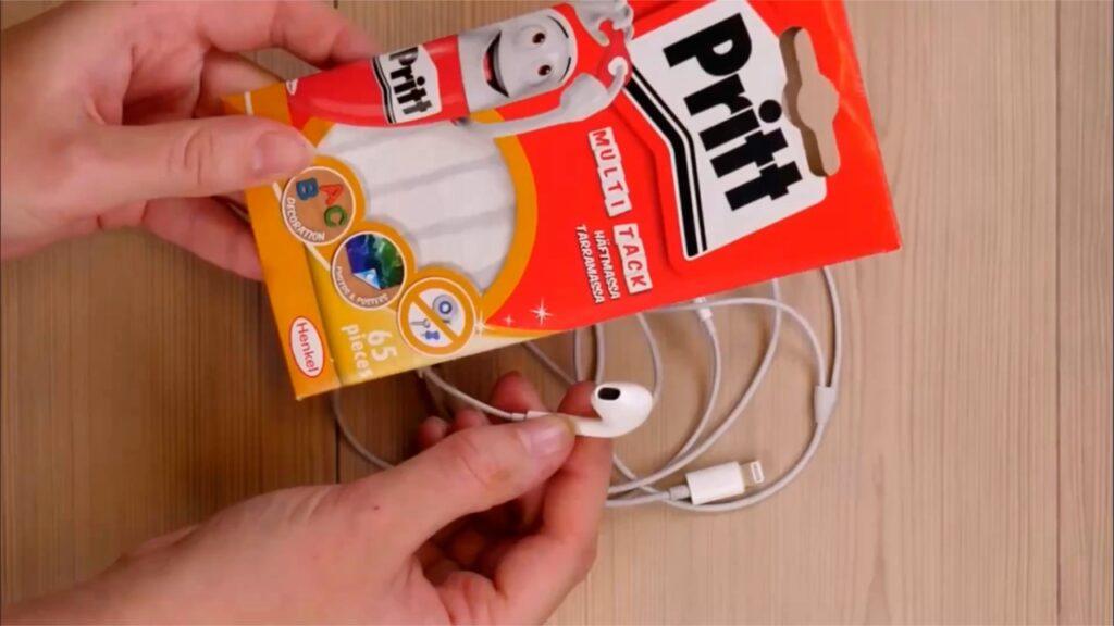 Bildet viser en person som holder en pakke med Pritt Multi Tack, et klebemiddel i form av små, hvite klumper. Pakken er rød og oransje, med en glad, animert Pritt-figur på forsiden. I tillegg til pakken holder personen også en hvit ørepropp, som er koblet til en hvit kabel med en Lightning-kontakt, mest sannsynlig til bruk med Apple-enheter. Øreproppene og Pritt-pakken ligger mot en lys, trefarget bakgrunn.