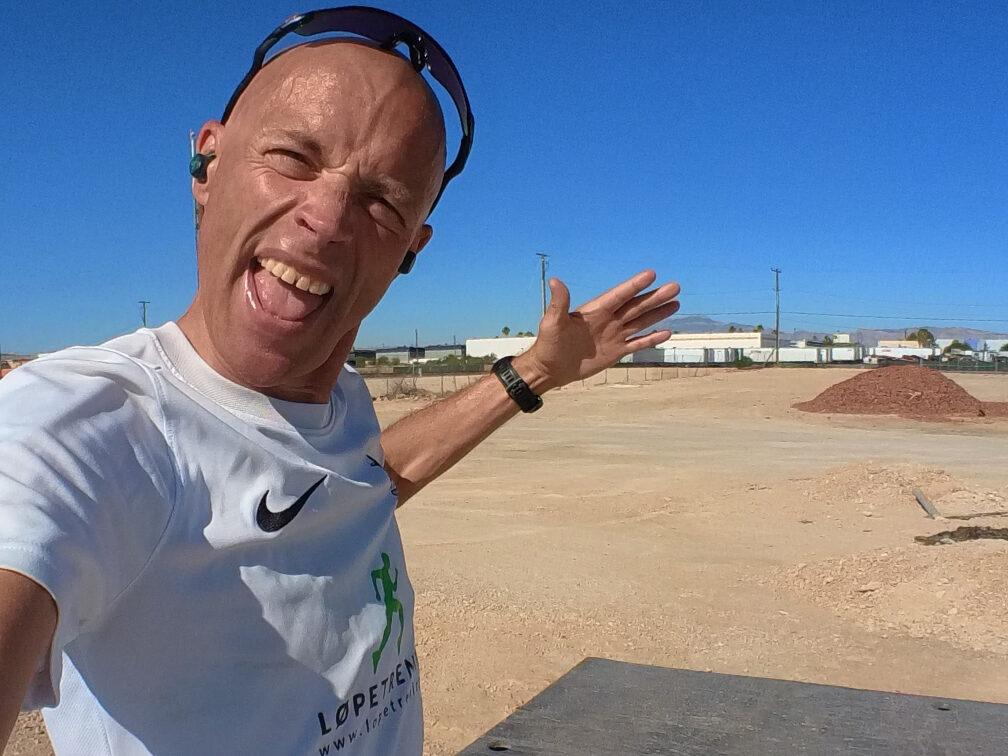 Bildet viser en entusiastisk mann som tar en selfie i et solrikt, ørkenlignende område. Han er iført en hvit treningsskjorte med "Løpetrening"-logoen og Nike-emblem på brystet. Mannen har solbriller på hodet og ser ut til å være i godt humør, med et bredt smil og utstrakte armer. I bakgrunnen ser man en tørr, åpen slette med noen få bygninger og jordhauger, og himmelen er knallblå uten skyer. Mannen har ørepropper i ørene, noe som tyder på at han lytter til musikk eller en podcast mens han er ute. Stemningen i bildet er energisk og positiv. Løpeeksperten Andreas Gossner fra Løpetrening.no deler sin treningsfilosofi.