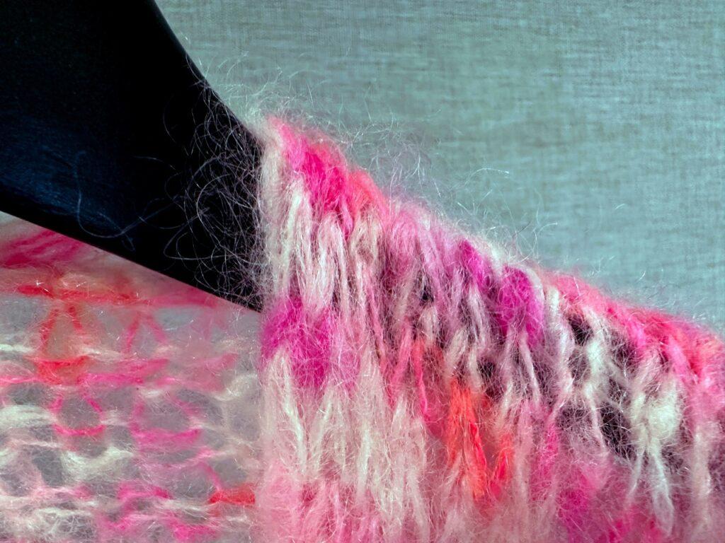 Bildet viser en strikket jakke laget av silk mohair-garn, kjent for sin luftige og myke tekstur, men også for å slippe løse fibre. Jakken har en fargerik blanding av rosa, lilla og kremhvit, med tydelige, bustete ulltråder som stikker ut. Den er drapert over en sort kleshenger, som skaper en kontrast mot de myke fibrene. Bakgrunnen er nøytral, noe som lar teksturen og fargene i garnet stå frem. Dette bildet illustrerer hvordan slike ullplagg kan røyte, spesielt når de er laget av mohair, og kan brukes i en artikkel om metoder for å minimere røyting.