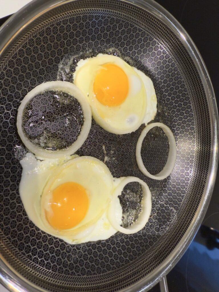 Et nærbilde av en hvit, hel egg plassert på en moderne stekepanne med en bikubemønstret non-stick-overflate. Over pannen ses en hånd med lange, røde negler som er i ferd med å slippe egget forsiktig ned. Pannen står på en blank induksjonstopp, og bakgrunnen har diffuse refleksjoner av lys. Bildet har et stilrent og minimalistisk preg, som fremhever kjøkkenredskaper og detaljer.