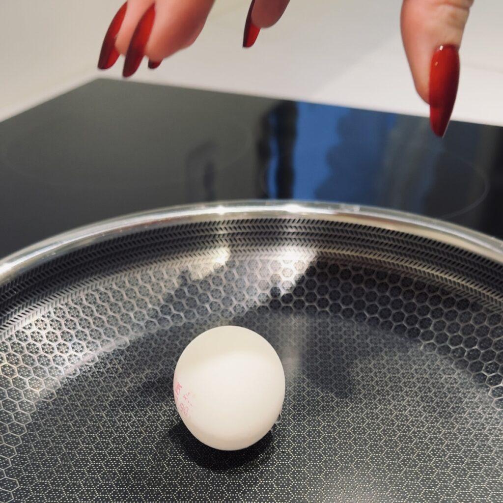 Et nærbilde av en hvit, hel egg plassert på en moderne stekepanne med en bikubemønstret non-stick-overflate. Over pannen ses en hånd med lange, røde negler som er i ferd med å slippe egget forsiktig ned. Pannen står på en blank induksjonstopp, og bakgrunnen har diffuse refleksjoner av lys. Bildet har et stilrent og minimalistisk preg, som fremhever kjøkkenredskaper og detaljer.