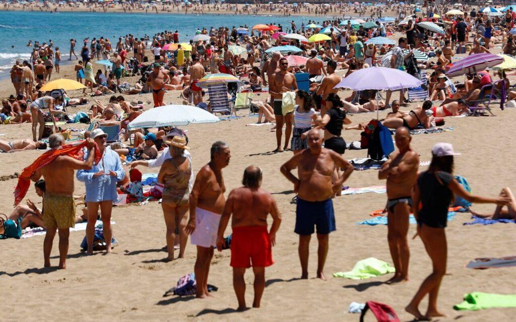 Bildet viser en travel sandstrand full av mennesker som nyter sol og varme. Stranden er tettpakket med solsenger, parasoller og strandutstyr. Mange mennesker står eller sitter i små grupper, mens andre ligger på håndklær og soler seg. Havet i bakgrunnen er blått, og vi kan se flere mennesker som bader. Langs kystlinjen skimtes bygninger, noe som tyder på at dette er en strand nær en by eller et populært turistområde, sannsynligvis et sted som Gran Canaria. Atmosfæren er livlig, og det ser ut til å være en varm og solrik dag.