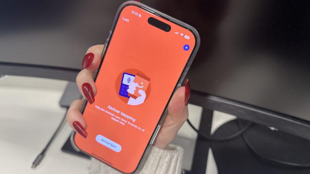 Nærbilde av en hånd med røde negler som holder en mobiltelefon. Skjermen viser Vipps-appen med en oransje bakgrunn og teksten 'Aktiver tæpping' for å aktivere kontaktløs betaling. En kortterminal og mobil-ikon vises i illustrasjonen på skjermen.