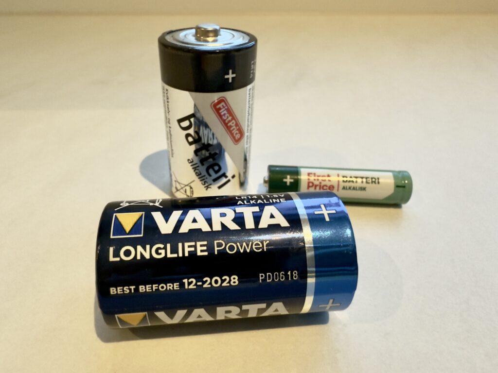 Tre forskjellige alkaliske batterier ligger på en lys overflate. Foran er et Varta Longlife Power D-batteri med en utløpsdato på desember 2028 tydelig synlig. Bak til venstre står et First Price alkaliskt C-batteri i sølvfarget emballasje, mens et grønt og hvitt First Price AAA-batteri ligger horisontalt til høyre. Batteriene illustrerer variasjon i størrelse og merke, og er oppstilt med god belysning for å fremheve detaljer i emballasjen.