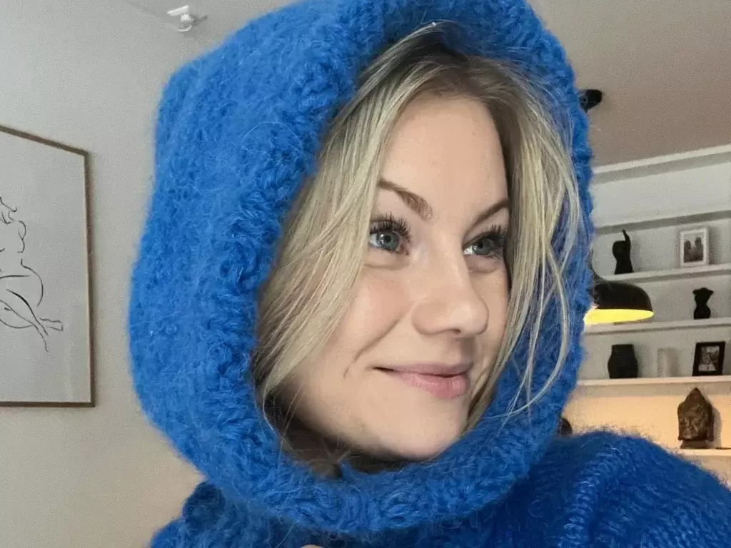En kvinne med blondt hår og blå øyne smiler og ser til siden. Hun har på seg en blå, strikket balaklava laget av mykt garn. Bakgrunnen viser en moderne stue med dekorative hyller.