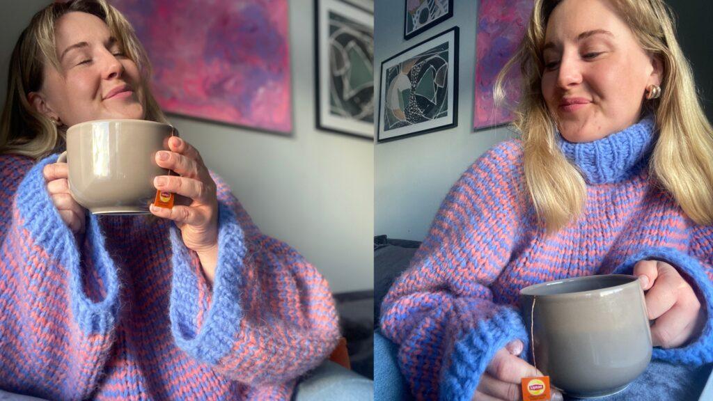 To bilder av en smilende kvinne i den håndstrikkede Cozy Stripes-genseren fra Vild Knitwear. Genseren er oversized med høy hals og strikket i brede, tofargede striper i lavendelblått og rosatoner. Hun holder en stor beige tekopp med Lipton-tepose, omgitt av moderne kunst på veggen i bakgrunnen. Stemningen er varm, avslappet og hjemmekoselig.