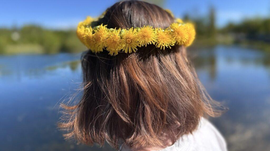 Bakhode av en person med skulderlangt, brunt hår iført en krans av gule løvetannblomster. I bakgrunnen skimtes et stille, blått vann med uskarpe trær langs bredden under en klar, blå himmel.