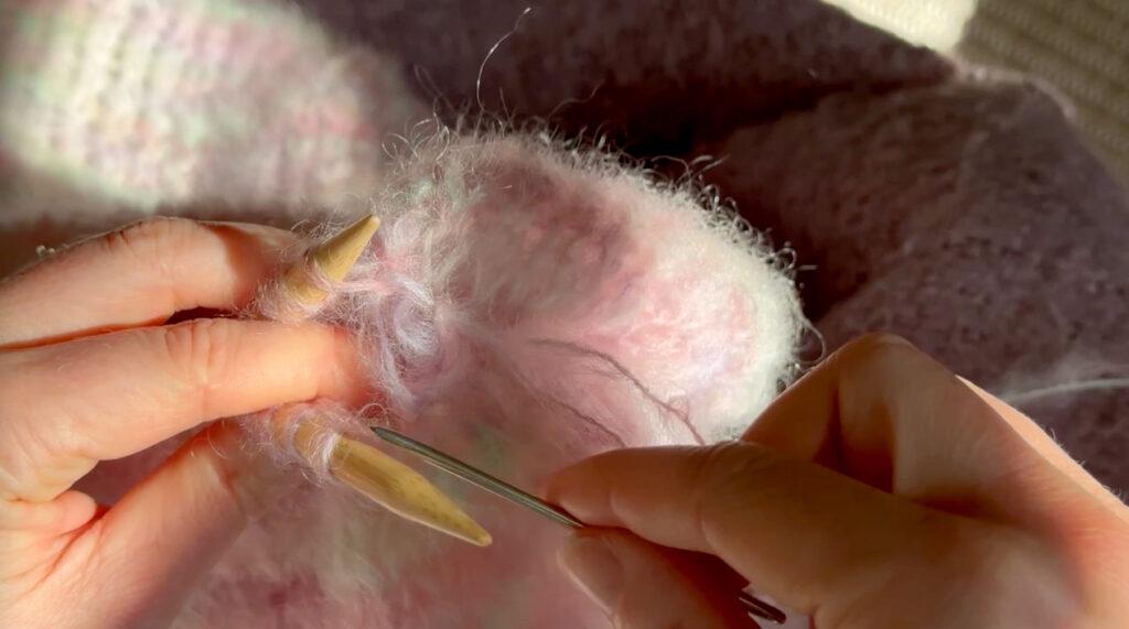 Nærbilde av teknikken “maske sammen” (kitchener stitch) som brukes til å veve sammen skuldersømmene på Sukkerspinnjakken. Bildet viser en ullnål som føres gjennom løkkene på to strikkepinner med mohairstrikk, i naturlig lys. Garnet er pastellfarget og mykt, og teknikken gir en usynlig søm som ser ut som glattstrikk.