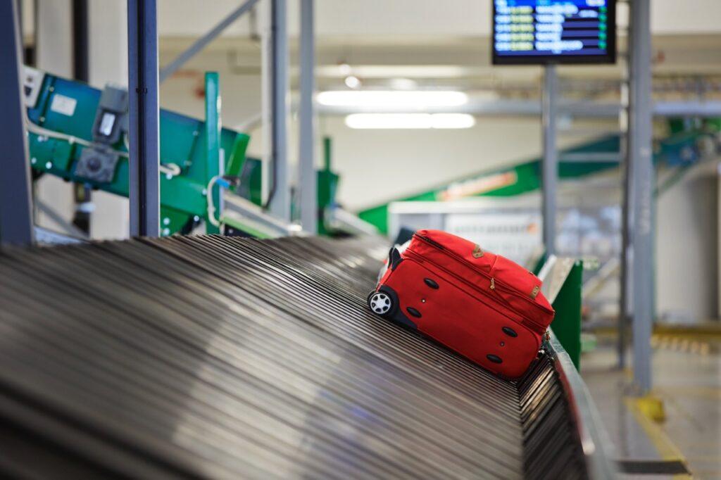 Bilde av rød koffert på rullebånd på flyplass. Vi gir deg tipset som sikrer at du får bagasjen din først på bagasjebåndet!