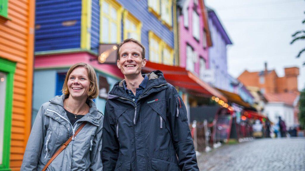 Smilende mann og kvinne i fargegaten i Stavanger. Full av fargerike trehus, gul, blå, lilla, rosa, grønn og oransje i bakgrunnen.
