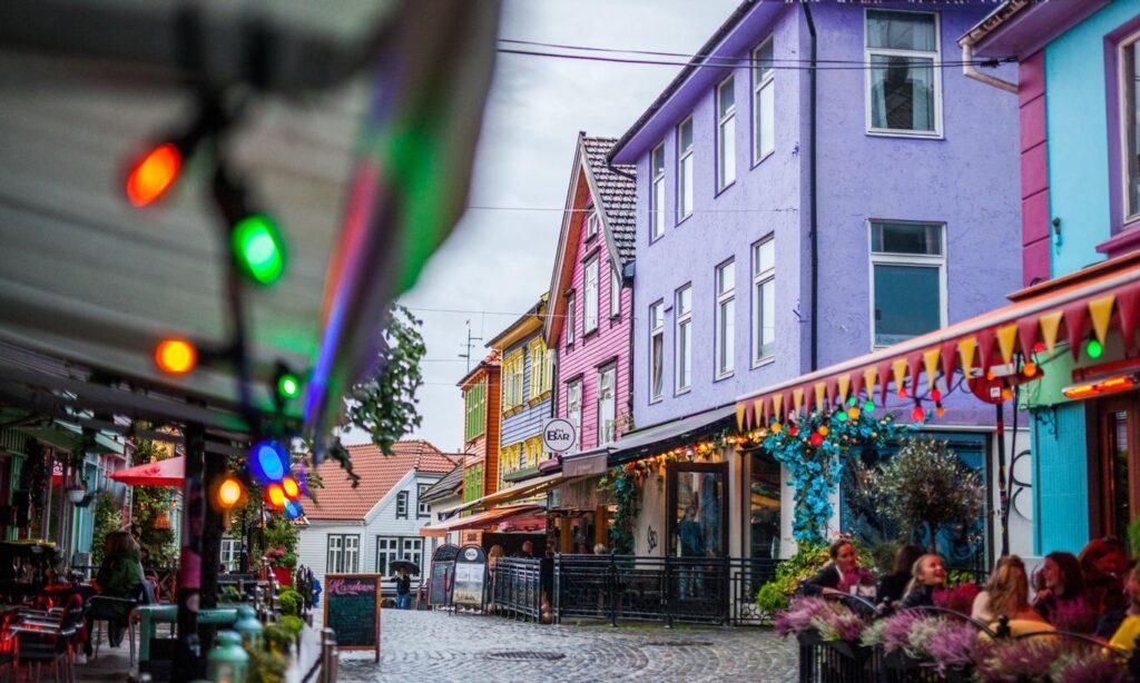 Folk på kafé i fargegaten i Stavanger. Full av fargerike trehus, gul, blå, lilla, rosa, grønn og oransje, med lyslenke i front.