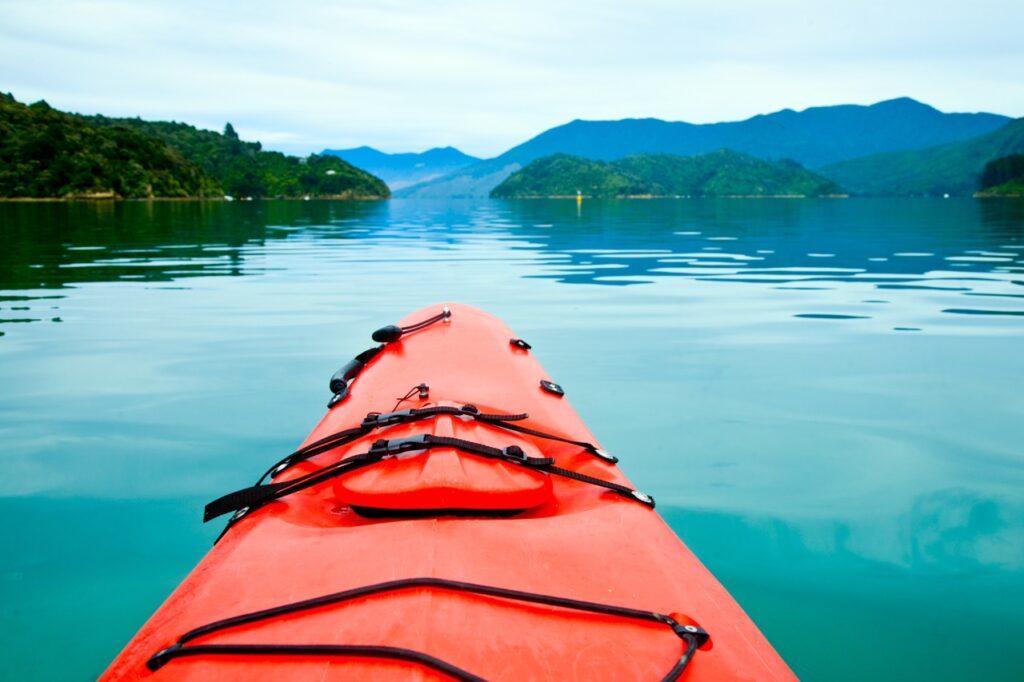Rød kajakk på blått stille vann med fjell i bakgrunn. Vi gir deg de beste rådene slik at du finner riktig kajakk til ditt bruk, enten du er nybegynner eller erfaren padler.