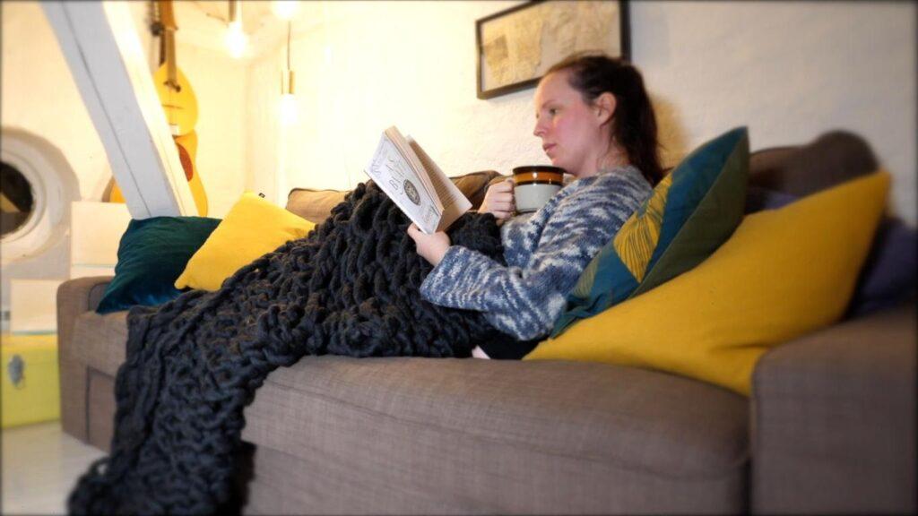 Kvinne sitter i sofa og leser med et teppe strikket med armene.
