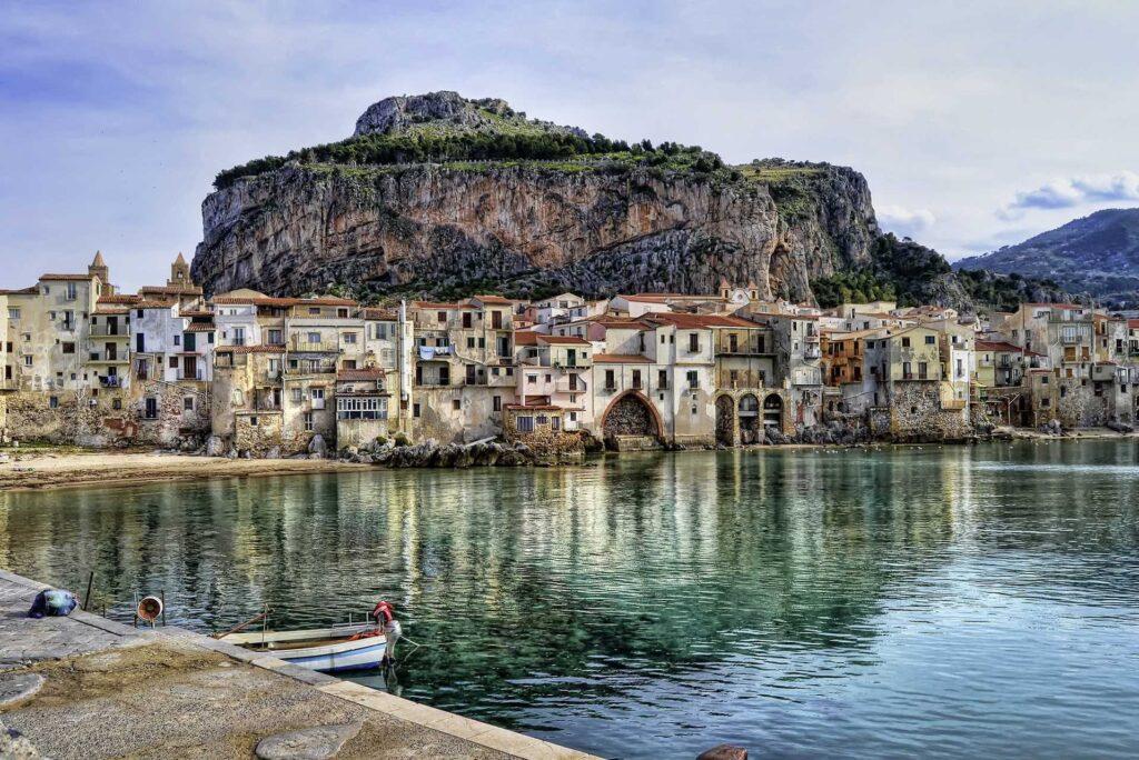 Sommerferie og sydentur til denne spennende landsbyen på Sicilia. Bilde av blått hav med en gammel landsby og fjell i bakgrunnen.