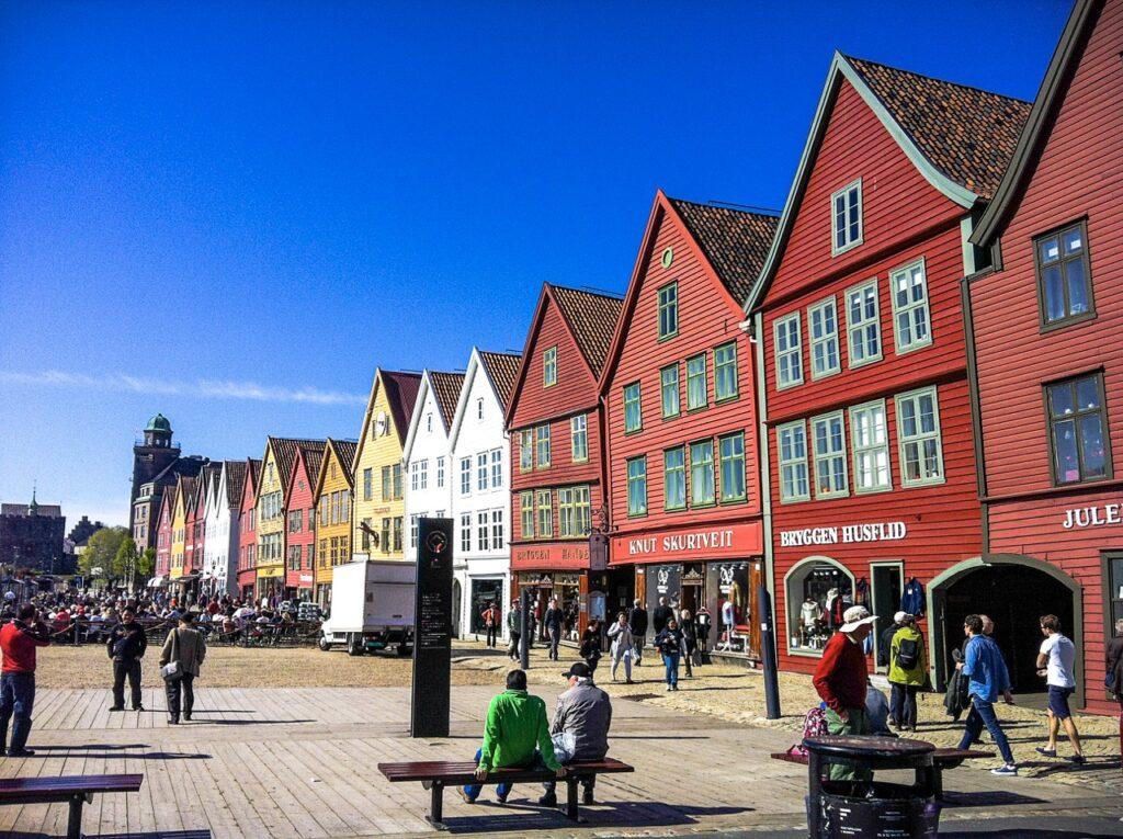 Bryggen i Bergen, med folk som sitter på benk og slikker sol en nydelig solskinnsdag. Fargerike trehus.