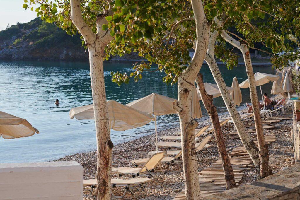 Bilde av strand på Samos i Hellas, med parasoll og solsenger, trær og badende person.