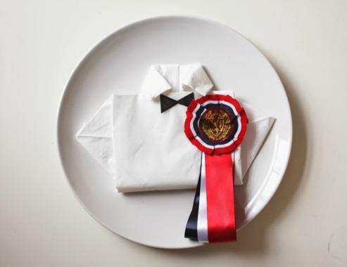 Bilde av serviett brettet som en skjorte, på en tallerken, med 17. mai-sløyfe til nasjonaldagen.