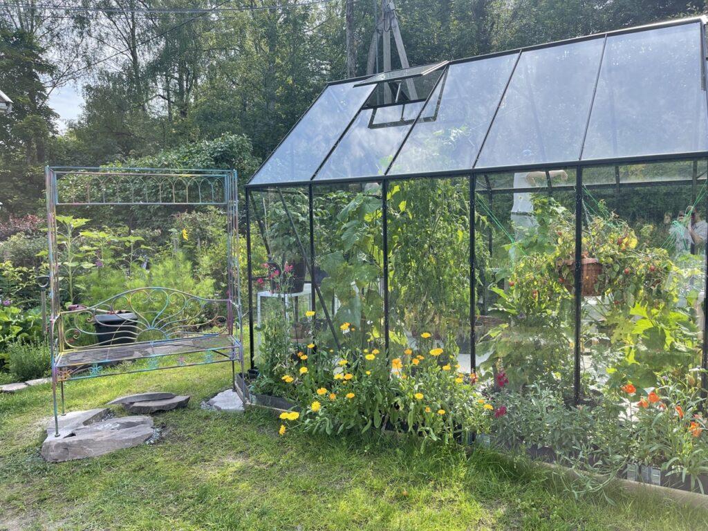 Bilde av drivhus med plantekasse, blomster og stålhylle utenfor, i en frodig hage. 