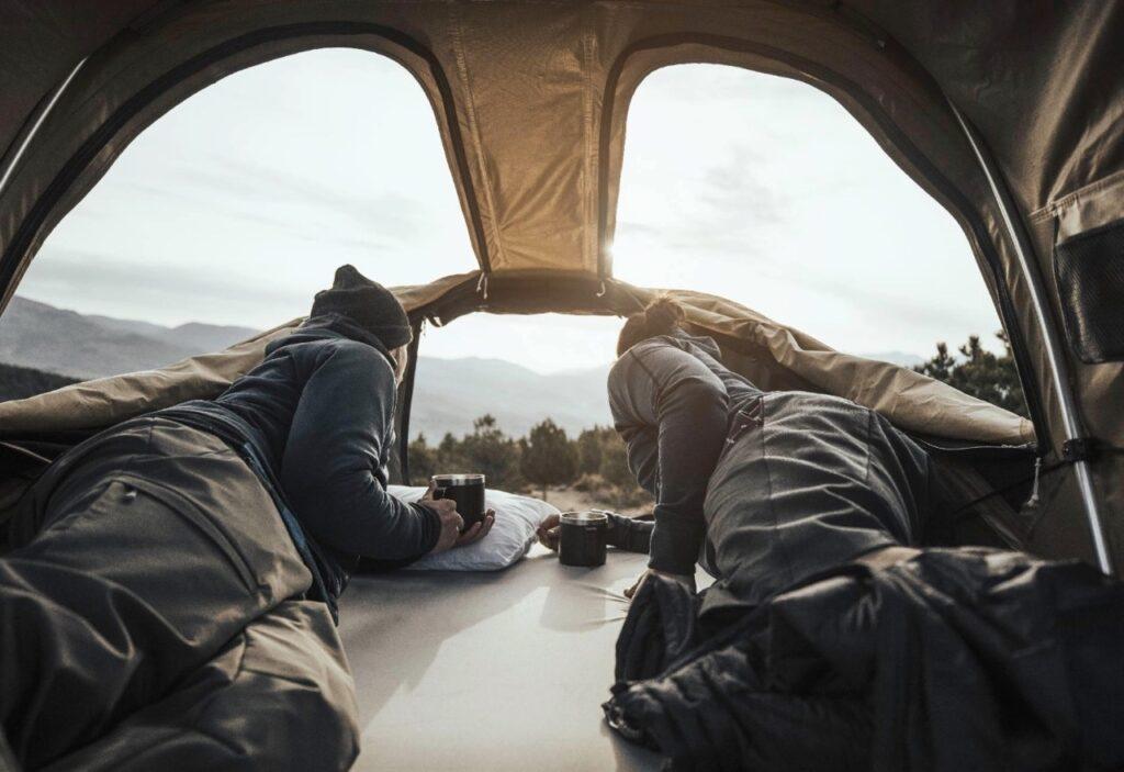 Bilde av to personer som ligger i et beige taktelt på bil og titter ut i naturen. Her får du  