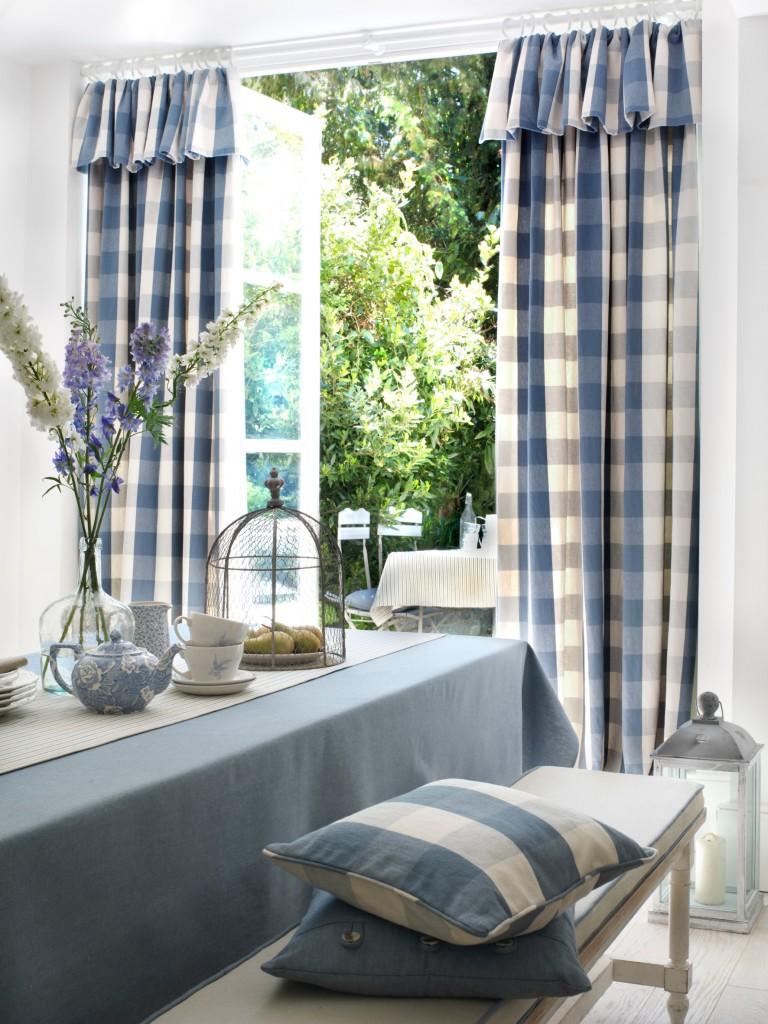 Blårutete gardiner med kappe ut mot natur og hage med spisebord med blå og hvit duk foran.