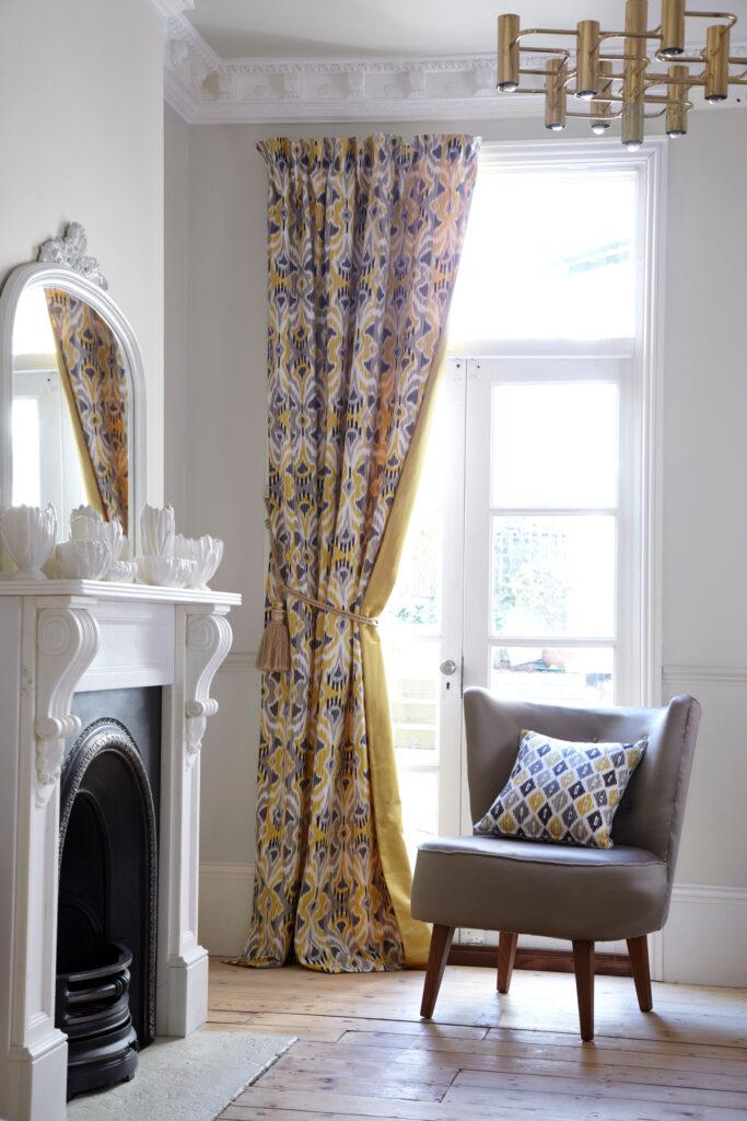 Gule gardiner med mønster og gult for i hvit stue bak stol.