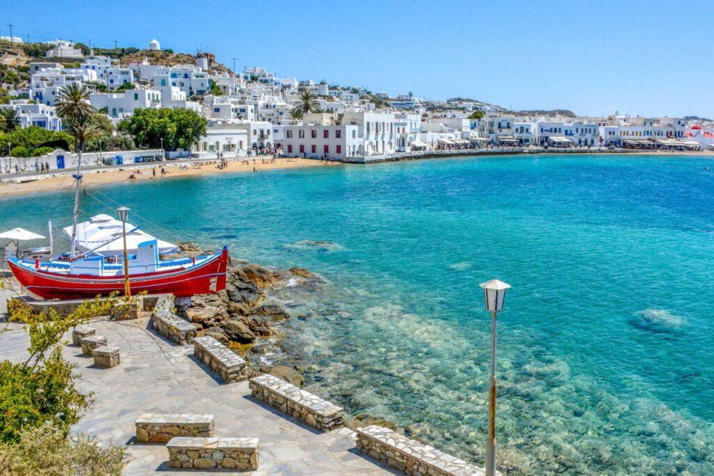 Nå lanseres det også reiser til Mykonos i Apollo sine kombinasjonreiser til Hellas. Her er en strand og havn med båt samt strandpromenade avbildet fra den vakre øya.