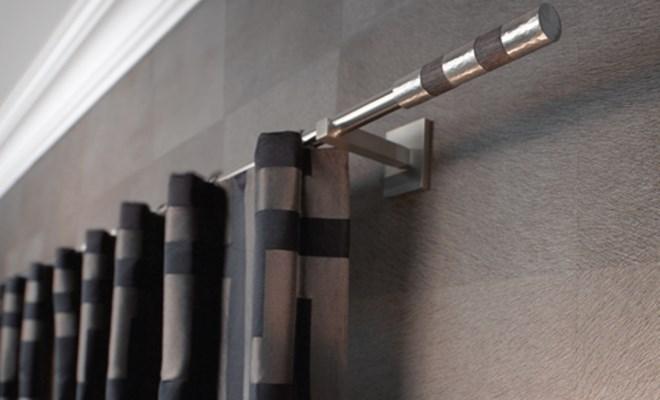 Grå gardiner med grafisk mønster på gardinstang i sølv mot grå tapet på vegg.