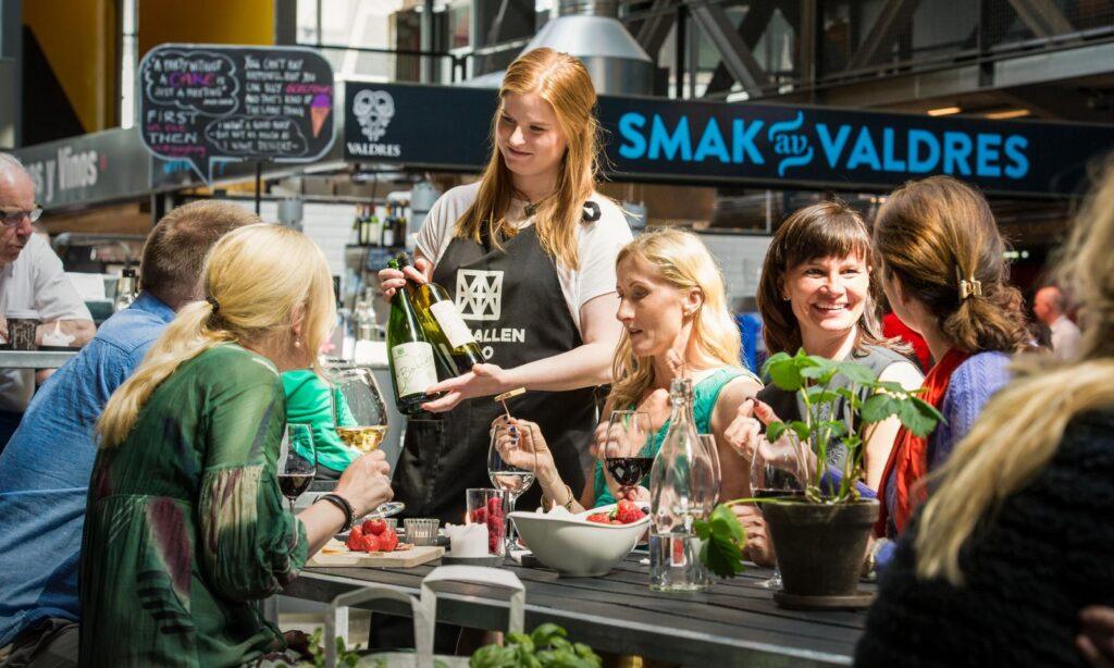 Bilde av servitør og kvinner ved bord med mat på Mathallen på Vulkan-området i Oslo.