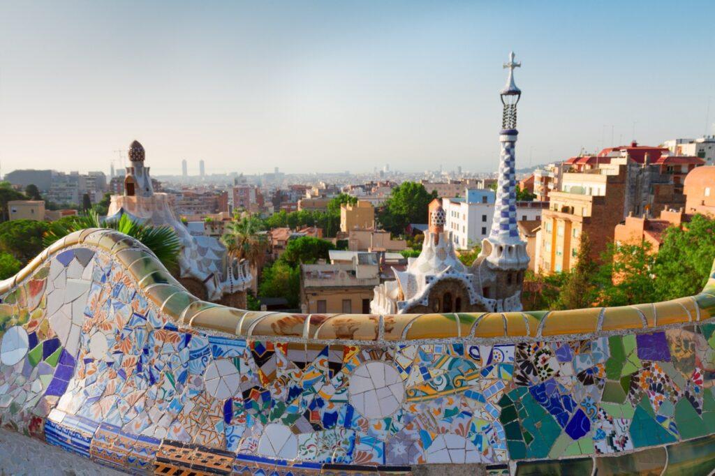 Bilde fra Park Guell i Barcelona hvor du ser byen i bakgrunnen og fargerike mosaikk kunst på gjerder langs en vei. Vi har samlet fem ting du må se og oppleve i Barcelona.