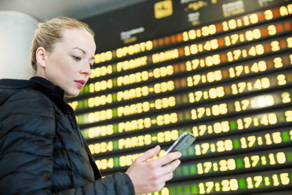 kvinne sjekker smarttelefonen sin mens hun står ved flight information-boardet.