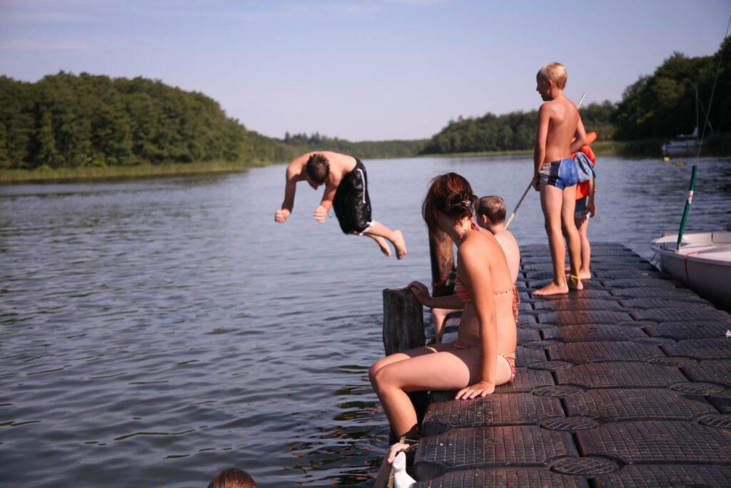 fem barn som bader i en innsjø i sommerferien. en stuper ut i vannet og de fire andre barna sitter på brygga. Dette er en sommerferie-aktivitet som er gratis.