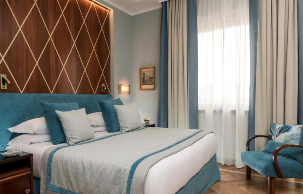 Hotellrom i 1930-talls stil med dobbeltseng og store vinduer med lyseblå og beige gardiner og sengeteppe.