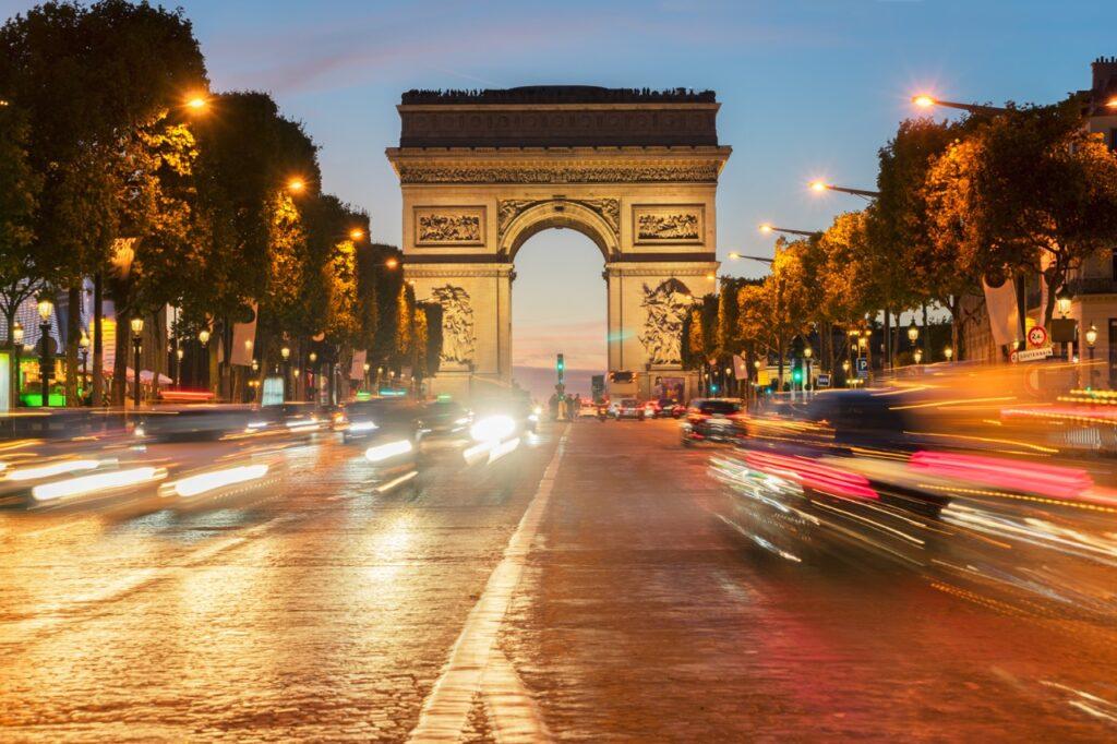 Paris - triumfbuen/ Arc de Triomphe en kveld med gatelys og trafikk.