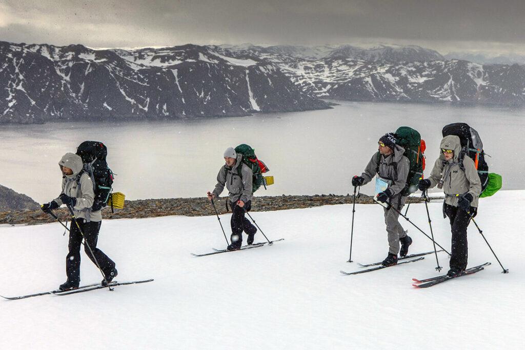 Bilde av deltakere i 71 grader nord som går på ski i snøvær ved en fjord og på et fjell. Personene har på seg grå skalljakker og tursekker, noen solbriller og luer og hansker. Vi forteller hvilke klær deltakerne i 71 grader nord har på seg i årets (2023) og programmets 15. sesong av programmet.