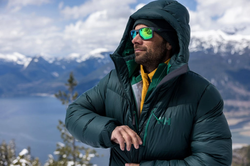 Bilde av mann i grønn skaljakke med solbriller og sekk på toppen av et fjell med en fjord i bakgrunn. Vi forteller hvilke klær deltakerne i 71 grader nord har på seg i årets (2023) og programmets 15. sesong av programmet.