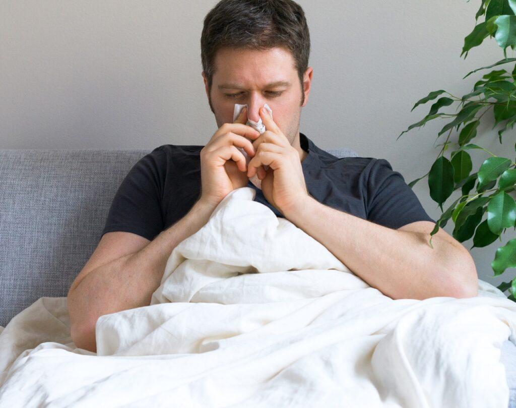 Bilde av syk mann med dyne i sofa, som snyter seg. Dette er spørsmålene du man stille deg om influensa 2022. 