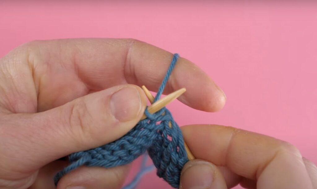 Bilde av hvordan strikke vrange masker med blått garn, strikkepinner i tre og rosa bakgrunn