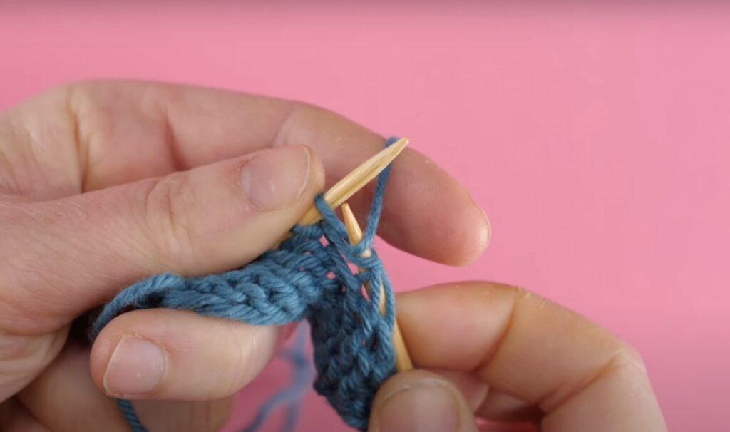 Bilde av hvordan strikke vrange masker med blått garn, strikkepinner i tre og rosa bakgrunn