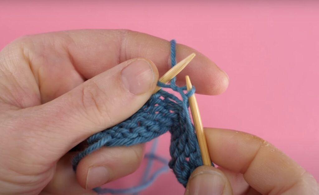 Bilde av hvordan strikke vrang med blått garn, strikkepinner i tre og rosa bakgrunn