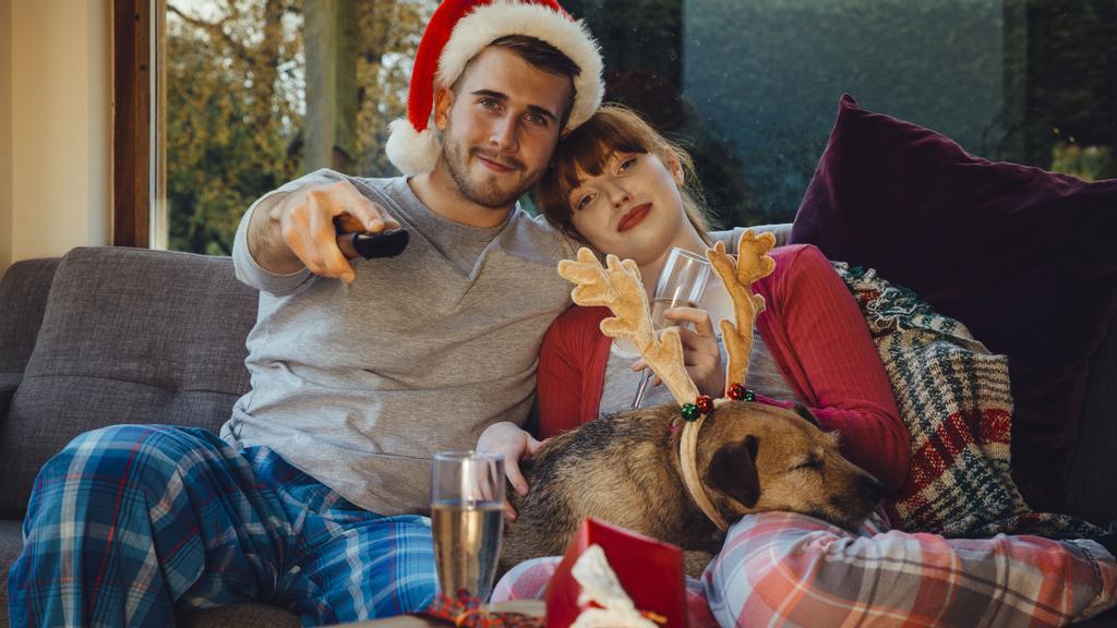 Mann med nisselue, dame og hund som sitter sammen i sofaen i pysj for å se på julefilm.