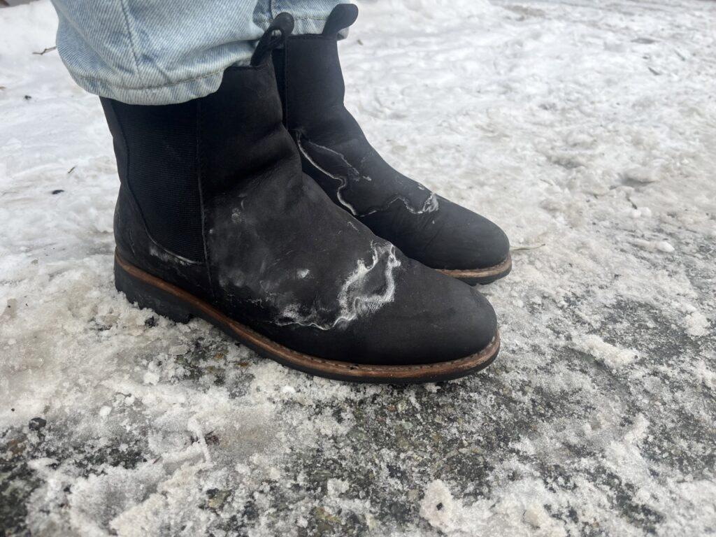 Bilde av sko med saltflekker etter vinterens veisalting. Skoene er svarte skinnsko og de har en veldig tydelig saltrand. Personen som har skoene på har også på seg en dongeribukse og står ute på en snødekket asfalt. Vi forteller hvordan du selv kan fjerne salt fra skoene dine.